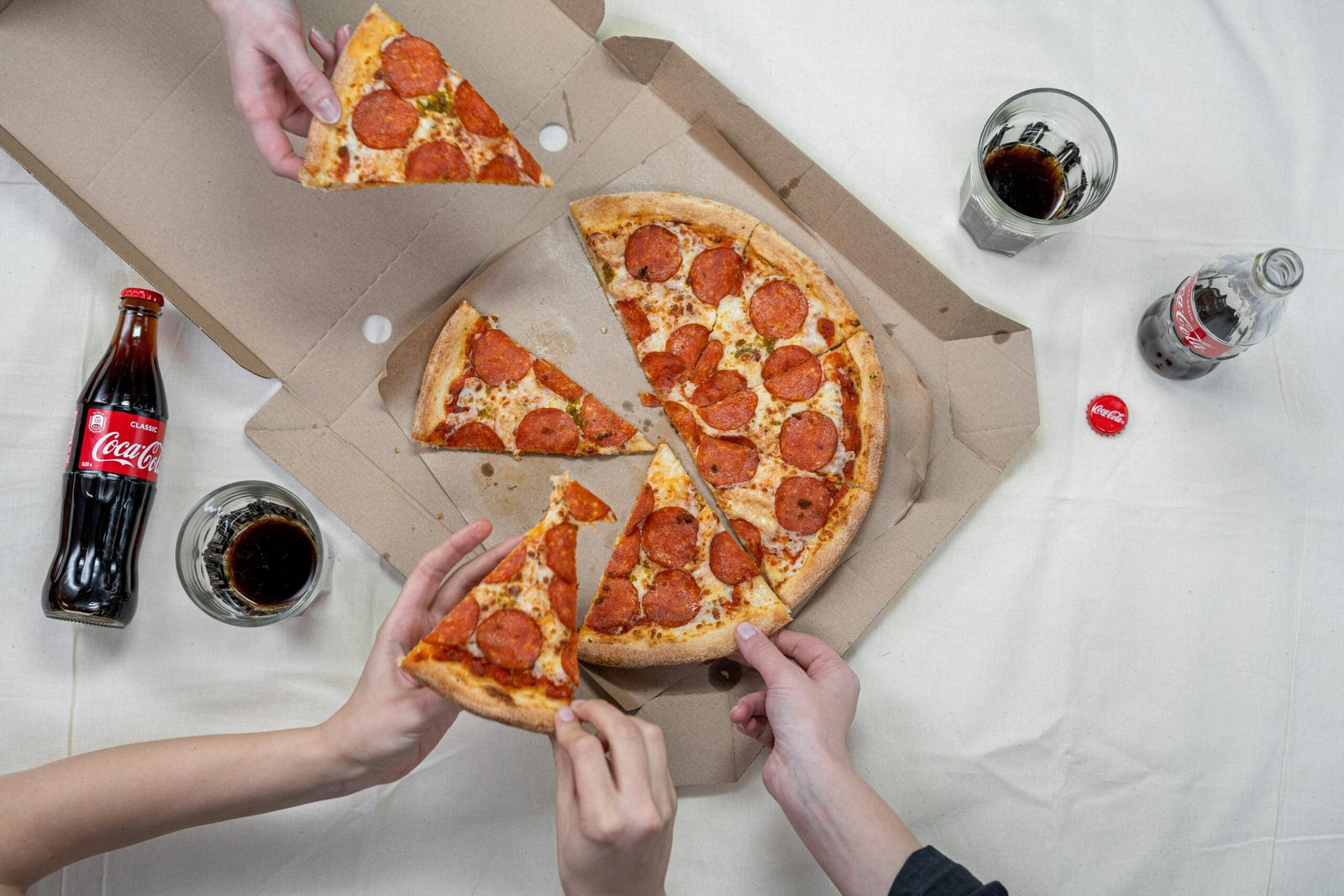 Top view of friends sharing a pepperoni pizza with Coca-Cola drinks on a white tablecloth.